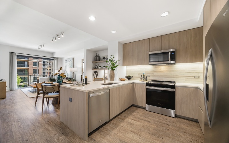 apartment kitchen with u-shaped counter, light flat wood cabinetry, stainless steel appliances, and white stone counters